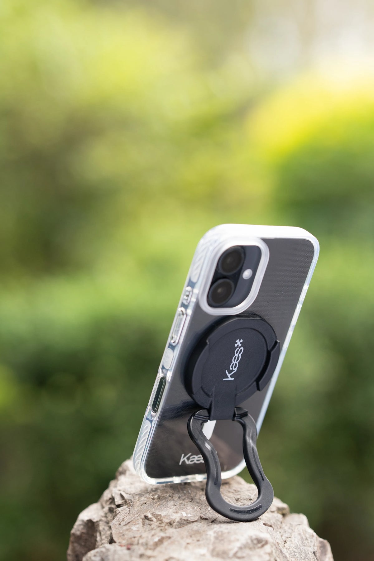 A clear and black protective Kaes iPhone case with a built-in black magnetic Perch Kaes grip and stand, shown on a smartphone resting on a rock against a blurred green outdoor background. The brand name 'Kaes' is visible on the ring and near the bottom of the case.