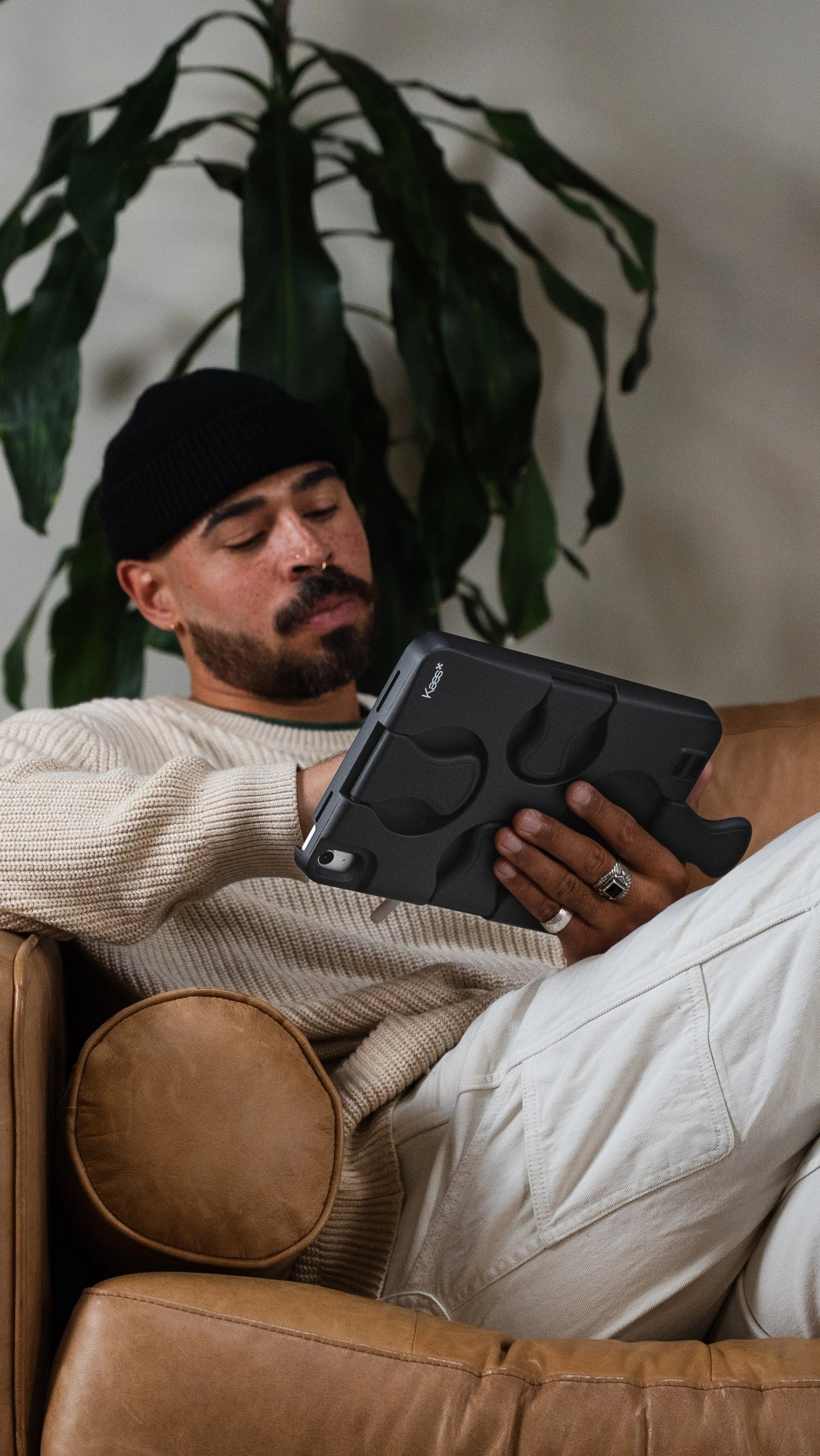 Man sitting on a brown leather couch holding a tablet with a plant in the background