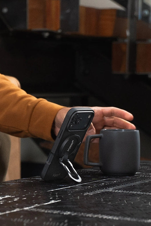 Person holding a black mug with a phone case attached, sitting at a table.