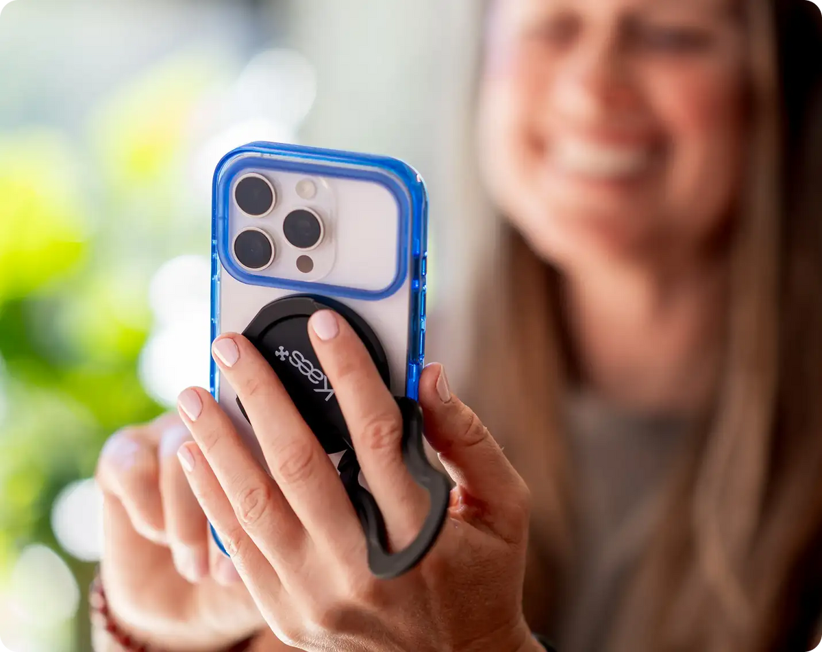 A close-up lifestyle shot of a person holding a smartphone with a blue and black Kees Perch and iPhone case against a blurred outdoor background.